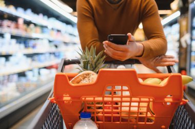 Alışveriş arabasını iten bir adam markette akıllı telefon kullanarak modern bir alışveriş deneyiminin tadını çıkarıyor.