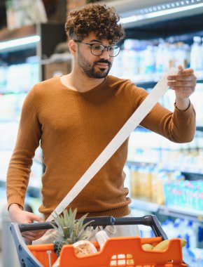 Müşteri uzun bir market fişini kontrol ederken süpermarket koridorlarında sepetle dolaşırken, harcamaları yönetirken ve akıllıca bütçe hesaplarken.