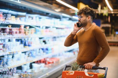 Müşteri akıllı telefonuyla konuşuyor, alışveriş arabasını itiyor ve markette yiyecek seçiyor.