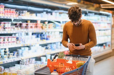 Müşteri, akıllı telefonuyla alışveriş arabasını parlak ve düzenli bir süpermarket koridorunda iterken market listesi uygulaması kullanıyor.