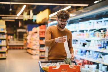 Genç adam süpermarkette alışveriş yaparken market fişini kontrol ediyor. Bütçeyi aşmadığından emin oluyor.