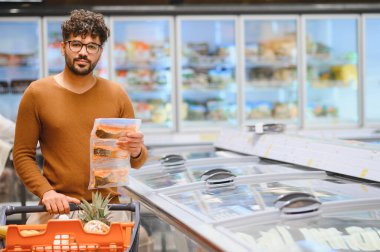 Süpermarkette dondurulmuş gıda reyonunda yiyecek seçen, donmuş somon paketleri tutan ve alışveriş arabası satan bir adam.