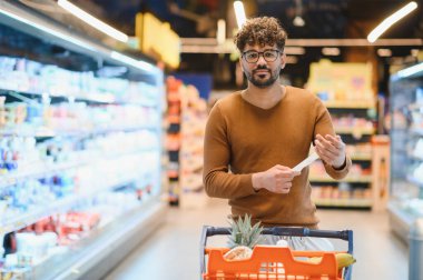 Müşteri alışveriş listesini okurken dolu bir alışveriş arabasını kalabalık bir süpermarketin koridorlarında itiyor.