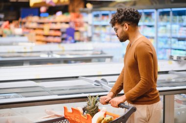 Genç Arap adam alışveriş arabasını itiyor ve dondurulmuş ürünleri süpermarket buzdolabı bölümünden seçiyor.