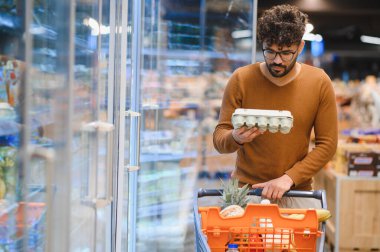 Müşteri marketin dondurulmuş bölümünde yumurtaları kontrol ediyor, alışveriş arabasını taze ürünlerle itiyor.
