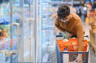 Arap adam market alışverişi yapıyor, alışveriş arabası itiyor ve dondurulmuş yiyecekleri süpermarket buzdolabı bölümünde seçiyor.