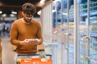 Müşteri markette alışveriş yaparken yiyecek etiketi okuyor, el arabası itiyor ve dondurulmuş bir ürünün malzemelerini kontrol ediyor.