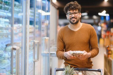 Bir süpermarkette market alışverişi yapan, dondurulmuş yiyecekleri elinde tutan ve alışveriş arabasını koridorlara iten bir müşteri.