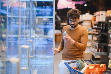 Odaklanmış Arap adam süpermarket buzdolabı koridorunda alışveriş yaparken su şişesi etiketini kontrol ediyor, bilinçli tüketici tercihleri yapıyor