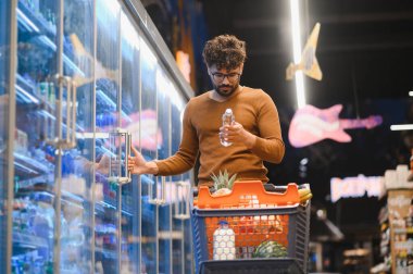 Genç adam market alışverişi yapıyor, buzdolabının kapısını açıyor ve süpermarkette su şişesini kontrol ediyor.