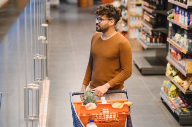 Genç Arap bir adam alışveriş arabasını iterken market koridorunda yiyecek ve taze yiyecek seçiyor.