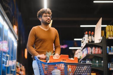 Genç Arap bir adam alışveriş arabasını iterken açık bir süpermarket koridorunda taze yiyecekler seçiyor ve uygun bir alışveriş deneyiminin tadını çıkarıyor.
