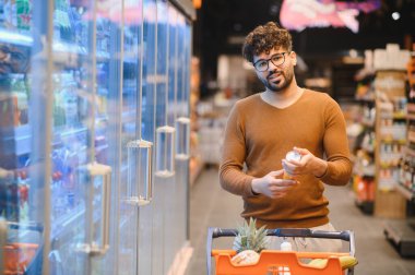 Genç Arap adam bakkaliye alışverişi, marketin buzdolabı bölümünden konserve ürünleri seçiyor, alışveriş arabası itiyor.