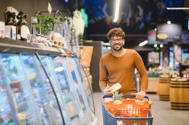 Genç bir Arap süpermarketten alışveriş arabası alırken elinde bir parça peynir ve gülümsüyor.