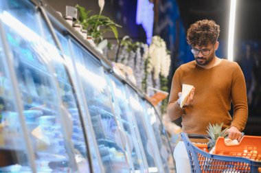 Müşteri marketin soğutulmuş bölümünden peynir seçiyor, bir parça peynir tutuyor ve alışveriş arabasını itiyor.