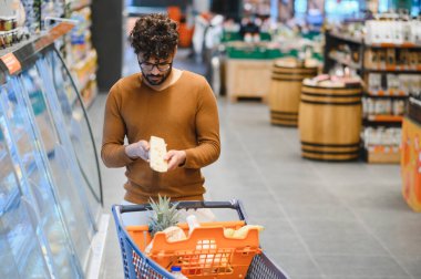 Müşteri, market alışverişi sırasında bir süpermarketin koridorlarında gezinirken peynir dilimindeki etiketi inceliyor.