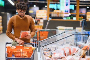 Genç bir Arap süpermarkette paketlenmiş eti dikkatlice seçiyor, alışveriş arabasını itiyor.