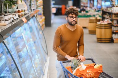 Müşteri, modern bir süpermarkette alışveriş arabasını itiyor, taze ürünlerle dolu parlak ışıklı raflardan sebze seçiyor.