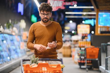 Akıllı telefondan alışveriş listesini kontrol eden bir adam, bir alışveriş arabasını parlak ve düzenli bir süpermarket koridorundan geçirirken,