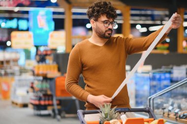 Genç adam süpermarkette alışveriş yaparken çok uzun bir market fişini dikkatle inceliyor ve artan gıda maliyetleri hakkındaki endişelerini dile getiriyor.