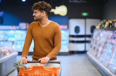 Genç Arap bir adam süpermarket koridorunda alışveriş arabasını itiyor, taze yiyecek arıyor ve sağlıklı yiyecek seçeneklerini seçiyor.