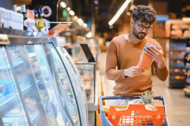 Genç adam marketin buzdolabı reyonunda alışveriş yaparken yiyecek etiketini dikkatlice okuyor.