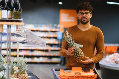 Markette ananas tutan, alışveriş arabasıyla yürüyen genç bir adam.