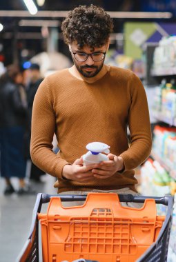 Genç adam bir marketteki ürünle ilgili malzemeleri ve besin bilgilerini dikkatlice kontrol ediyor, alışverişleri hakkında bilgilendirilmiş seçimler yapıyor.