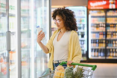 Genç bir kadın marketin soğutuculu bölümünde süt şişesi malzemelerini kontrol ediyor, içi taze ürünlerle dolu bir alışveriş arabasını itiyor.