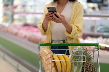 Müşteri akıllı telefon uygulaması kullanıyor ve markette içi taze ürün ve sebze dolu bir alışveriş arabasını ittiriyor.