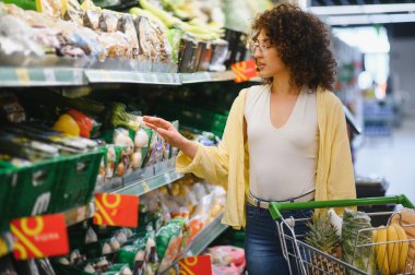 Raftan taze ürün seçen genç bir kadın süpermarket koridorunda meyve ve sebze dolu alışveriş arabalarını itiyor.