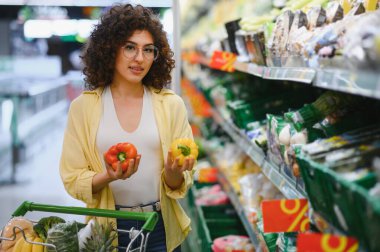 Müşteri, hareketli bir süpermarket koridorunda taze ürün alırken canlı kırmızı ve sarı dolma biberleri karşılaştırıyor