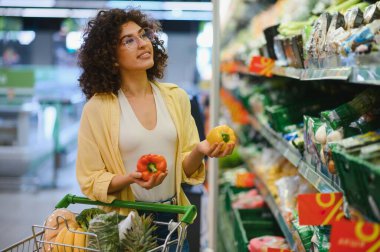 Müşteri, süpermarket koridorunda alışveriş yaparken canlı sarı ve kırmızı dolma biberleri karşılaştırıyor. Sağlıklı bir yemek için taze ürünleri dikkatlice seçiyor.