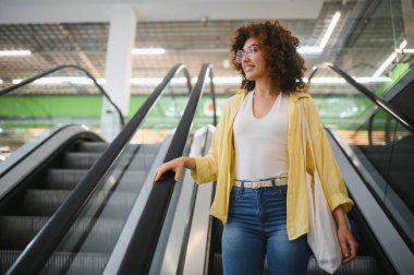 Bir markette yürüyen merdivene tekrar kullanılabilir bir alışveriş torbasıyla çıkıp gülümseyen ve başka tarafa bakan genç bir kadın.