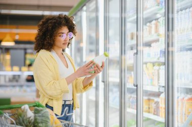 Sağlıklı seçeneklerini düşünerek markette alışveriş yaparken buzdolabı bölümünden süt şişesi seçen genç bir kadın.