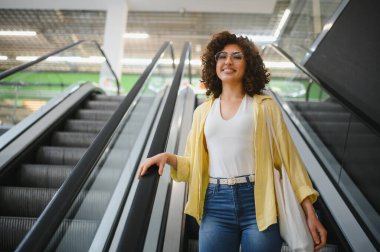 Yeniden kullanılabilir bir alışveriş çantası taşıyan genç bir kadın, parlak ve modern bir markette yürüyen merdivene tırmanıyor, alışveriş deneyiminin tadını çıkarıyor.