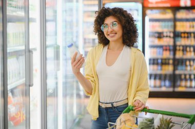 Süpermarkette buzdolabında alışveriş yaparken elinde süt şişesiyle gülümseyen kadın sağlıklı yaşam tarzı için taze süt ürünleri seçiyor.