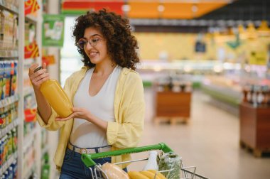 Markette meyve suyu taşıyan, gülümseyen ve alışveriş arabasını iten genç bir kadın.