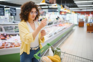 Alışveriş listesini kontrol eden genç bir kadın markette içi alışveriş arabasıyla geziyor.