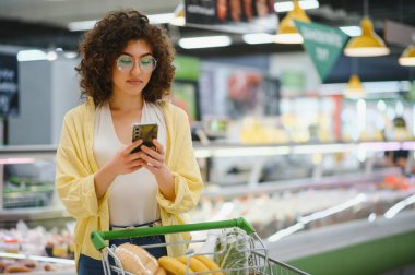 Akıllı telefonuyla meşgul olan genç bir kadın market koridorunda içi alışveriş arabasıyla koşturuyor.