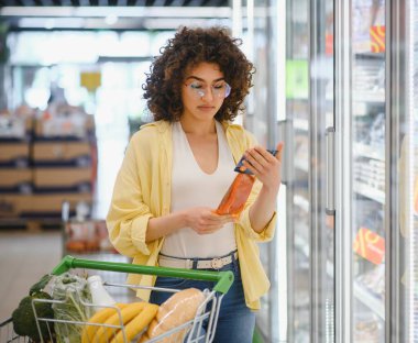 Genç bir kadın marketten alışveriş yaparken dondurulmuş gıda paketinin malzemelerini ve besin bilgilerini dikkatlice kontrol ediyor.