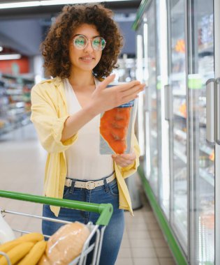 Market koridorunda alışveriş yaparken dondurulmuş somon filetosu seçen genç bir kadın.