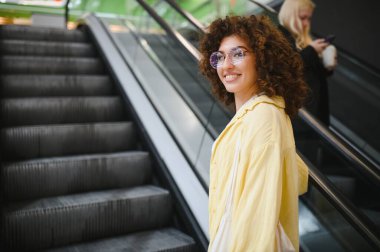 Kıvırcık saçlı ve gözlüklü genç bir kadın alışveriş merkezinde yürüyen merdivene çıkıyor.