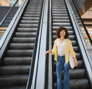 Markette yürüyen merdivenden inerken gülümseyen genç bir kadın, içi alışveriş torbalarıyla dolu bir poşet taşıyor.