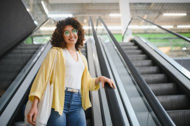 Bir süpermarkette yürüyen merdivene çıkan genç bir kadın, yeniden kullanılabilir bir alışveriş çantası taşıyor, modern ve çevre dostu bir alışveriş deneyimi yaşıyor.