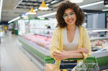 Gülümseyen genç bir kadın süpermarket koridorundaki zamanının tadını çıkarırken alışveriş arabasına yaslanıyor.