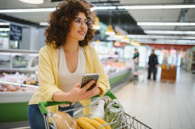 Müşteri akıllı telefonu karıştırırken marketin kalabalık koridorunda içi taze yiyeceklerle dolu bir alışveriş arabasını itiyor.