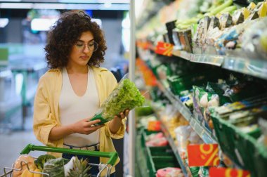 Marketten taze ürünler seçen, elinde marul ve sağlıklı yiyeceklerle alışveriş arabası tutan genç bir kadın.