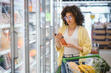 Müşteri, marketin dondurulmuş gıda bölümünde alışveriş yaparken farklı ürünlerin besin bilgilerini karşılaştıran bir akıllı telefon uygulaması kullanıyor.