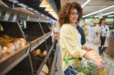 Market alışverişi yapan genç bir kadın, fırın bölümündeki raflardan bir somun ekmek topluyor.